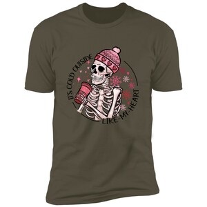 May include: Olive green t-shirt featuring a skeleton wearing a pink heart-patterned beanie and holding a red cup. The graphic includes the text "IT'S COLD OUTSIDE LIKE-MY-HEART" in a circular design with snowflakes.