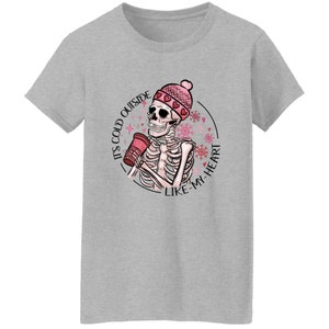 May include: A light gray t-shirt featuring a skeleton wearing a pink beanie with hearts and holding a red cup. The graphic includes the text "IT'S COLD OUTSIDE LIKE MY HEART" in a circular design with snowflakes.