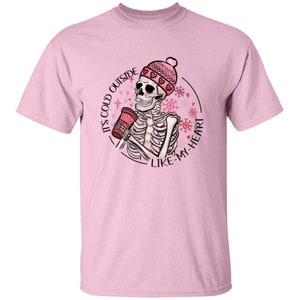May include: Light pink t-shirt featuring a skeleton wearing a pink beanie with hearts and holding a red cup. The shirt has the text "IT'S COLD OUTSIDE LIKE MY HEART" in a circular design with snowflakes.