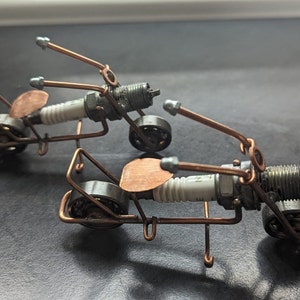以下が含まれることがあります： スパークプラグと銅線で作られたミニチュアバイク2台。 バイクは銅線フレーム、白いスパークプラグ、銅色のシートを備えています。
