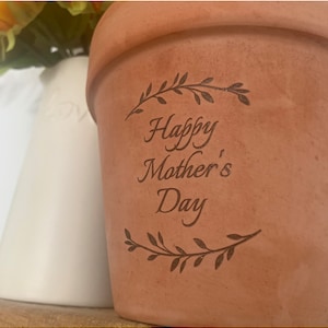 Könnte beinhalten: Ein Terrakotta-Blumentopf mit dem Text "Happy Mother's Day" auf der Seite. Der Topf ist mit einem einfachen Blumendesign verziert.