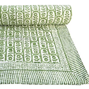 Peut inclure: Une courtepointe pliée présentant un motif répétitif de fleurs vertes dans des cadres ovales blancs sur fond blanc. La courtepointe possède une bordure avec un motif de vigne florale et un bord rayé, finie avec des points de piqûre blancs visibles.