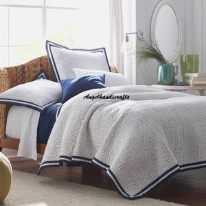 May include: A white and blue quilted bedspread with a navy blue trim. The bedspread is on a bed with a wicker headboard and a white pillow with a navy blue trim. There is a blue pillow on the bed and a green vase with a plant on a side table.