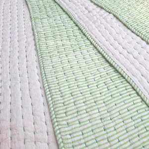 Op de afbeelding: Close-up van een wit en groen gestreept quilt. De quilt heeft een patroon van horizontale groene strepen op een witte achtergrond. De stiksels zijn zichtbaar en geven de stof textuur. Het ontwerp is eenvoudig en modern.