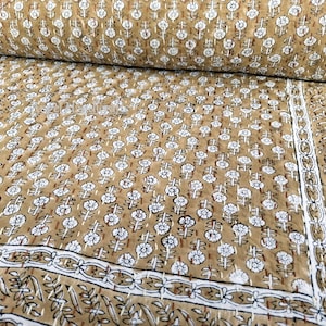 Op de afbeelding: Een quilt met een bloemenpatroon in bruin en wit. De quilt heeft een herhalend patroon van witte bloemen met bruine stelen en bladeren. De rand heeft een wit en bruin bloemmotief. De quilt is opgevouwen en opgerold.