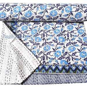 Op de afbeelding: Een opgerolde, witte quilt met een blauw bloemmotief en donkerblauwe ranken. De quilt is versierd met lichtblauwe bloemen en een donkerblauwe rand. De quilt is gemaakt van een zacht materiaal en is geschikt voor een bed of bank.