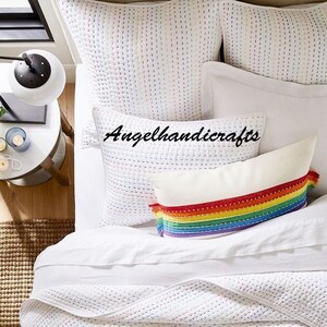 May include: White quilted bedding with a rainbow striped pillow with tassels. The pillow has a white background with a rainbow stripe pattern and white tassels on each end. The bedding is white with a subtle quilted pattern.