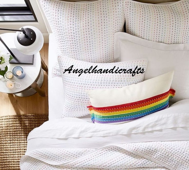 May include: White quilted bedding with a rainbow striped pillow with tassels. The pillow has a white background with a rainbow stripe pattern and white tassels on each end. The bedding is white with a subtle quilted pattern.