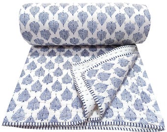 Puede incluir: Una colcha blanca con un patrón repetitivo de hojas azules y detalles de costura fina. El textil está parcialmente enrollado en la parte superior, revelando una esquina doblada con un borde de rayas azul oscuro que enmarca la pieza rectangular.