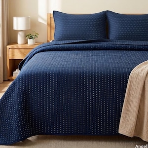 May include: A dark navy blue quilt set featuring a subtle white dashed vertical stitch pattern. The set includes two matching rectangular pillow shams and a large bedspread, complemented by a folded beige textured throw blanket draped on the side.
