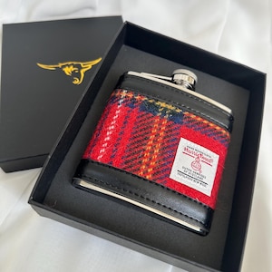 May include: A black box with a red and black flask inside. The flask is covered in a red, black, and yellow plaid fabric with a label that reads "Harris Tweed, Hand Woven Cloth, Outer Hebrides of Scotland, 100% Pure New Wool."