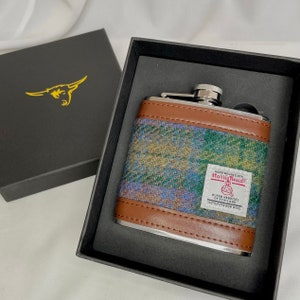 May include: A silver flask with a brown leather and tweed exterior. The flask is in a black box with a gold bull logo. The tweed is labeled "Harris Tweed" and "Outer Hebrides of Scotland".