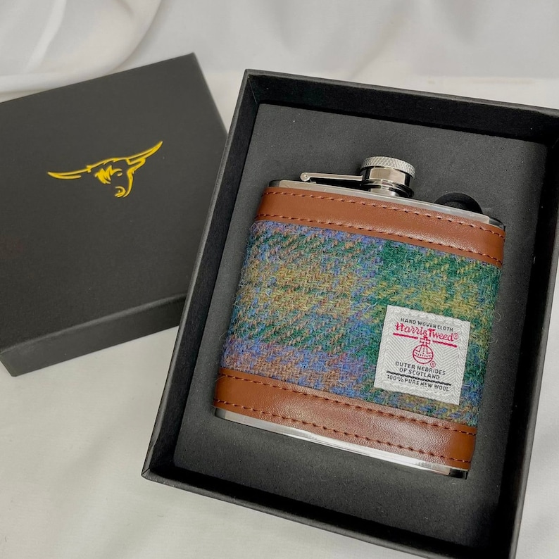 May include: A silver flask with a brown leather and tweed exterior. The flask is in a black box with a gold bull logo. The tweed is labeled "Harris Tweed" and "Outer Hebrides of Scotland".
