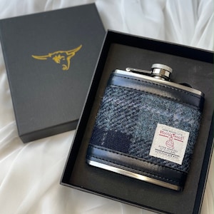 May include: A silver and black hip flask with a blue and white plaid pattern. The flask is made of stainless steel and has a leather-like finish. The flask is in a black box with a gold bull logo. The flask has a label that reads "Hand Woven Cloth Harris Tweed Outer Hebrides of Scotland 100% Pure New Wool"