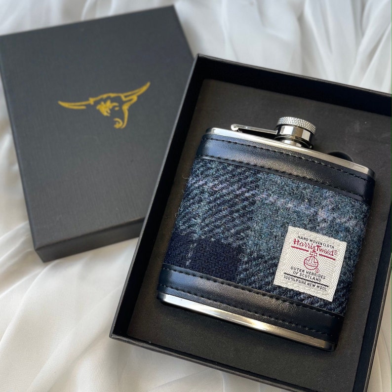 May include: A silver and black hip flask with a blue and white plaid pattern. The flask is made of stainless steel and has a leather-like finish. The flask is in a black box with a gold bull logo. The flask has a label that reads "Hand Woven Cloth Harris Tweed Outer Hebrides of Scotland 100% Pure New Wool"