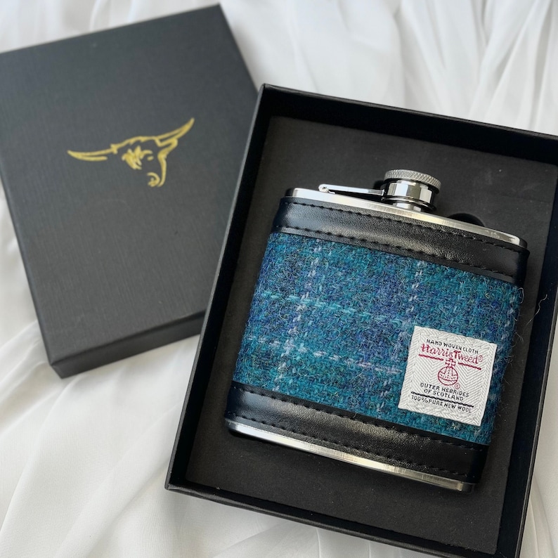 May include: A silver flask with a black leather trim and a blue and white tweed pattern. The flask has a label that reads "Hand Woven Cloth Harris Tweed Outer Hebrides of Scotland 100% Pure New Wool."