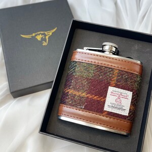 May include: A silver hip flask with a brown leather trim and a plaid pattern. The flask has a label that reads "Hand Woven Cloth Harris Tweed Outer Hebrides of Scotland 100% Pure New Wool".