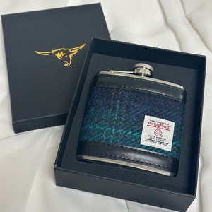 May include: A black and silver flask with a green, blue, and black tweed pattern. The flask has a label that reads "Handwoven Harris Tweed Outer Hebrides of Scotland 100% Pure New Wool".