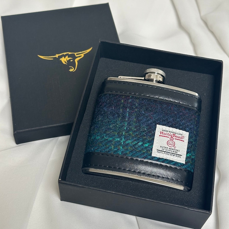 May include: A black and silver flask with a green, blue, and black tweed pattern. The flask has a label that reads "Handwoven Harris Tweed Outer Hebrides of Scotland 100% Pure New Wool".