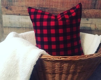 May include: A red and black buffalo plaid pillow rests in a woven wicker basket, alongside a fluffy white blanket. The pillow is square-shaped and the basket is brown. The background is a wooden wall.