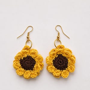 May include: A pair of dangle earrings featuring crocheted sunflower motifs. Each earring has a dark brown circular center surrounded by bright yellow petals, attached to a gold-toned metal hook and a small circular jump ring for movement.
