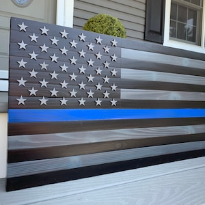 May include: A wooden American flag with a blue line across the middle. The flag is made of dark wood with a distressed finish. The stars are silver and the stripes are black and gray.