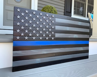 May include: A wooden American flag with a blue line across the middle. The flag is made of dark wood with a distressed finish. The stars are silver and the stripes are black and gray.