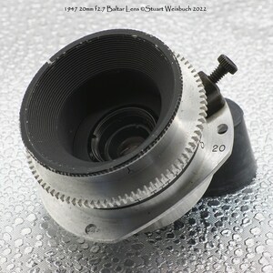 Puede incluir: Un objetivo de c&aacute;mara de color plateado con un borde negro y un anillo de enfoque negro. El objetivo tiene una distancia focal de 20 mm y una apertura de f/2,7. El objetivo est&aacute; etiquetado como "1947 20mm f2.7 Baltlar Lens &copy;Stuart Weisbuch 2022".