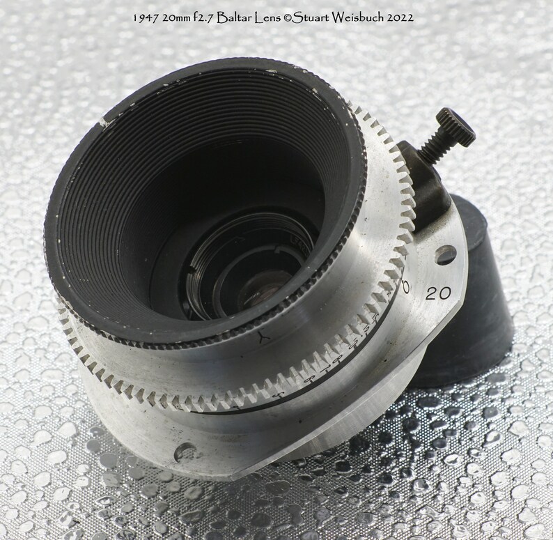 Puede incluir: Un objetivo de c&aacute;mara de color plateado con un borde negro y un anillo de enfoque negro. El objetivo tiene una distancia focal de 20 mm y una apertura de f/2,7. El objetivo est&aacute; etiquetado como "1947 20mm f2.7 Baltlar Lens &copy;Stuart Weisbuch 2022".
