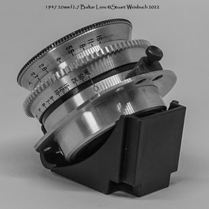 Puede incluir: Un objetivo de c&aacute;mara plateado con una capucha de lente negra. El objetivo tiene una escala de profundidad de campo marcada en pies y metros. El objetivo est&aacute; etiquetado como "Balta Lens".