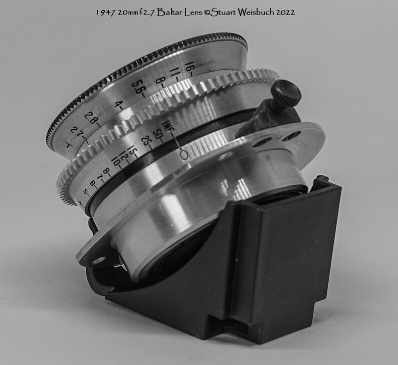 Puede incluir: Un objetivo de c&aacute;mara plateado con una capucha de lente negra. El objetivo tiene una escala de profundidad de campo marcada en pies y metros. El objetivo est&aacute; etiquetado como "Balta Lens".