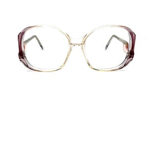 May include: Clear eyeglasses with a gradient frame, transitioning from a light beige to a deep burgundy at the temples. The lenses are large and round, with thin, silver-colored arms.