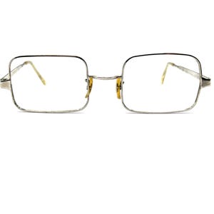 May include: Silver-framed eyeglasses with square lenses. The glasses have a thin, silver frame and temples, with a small bridge. The lenses are clear, and the overall design is simple and classic.