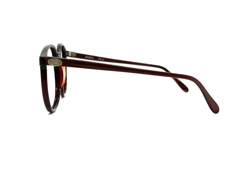 May include: A pair of brown and black round eyeglasses with a silver accent on the side. The glasses have the text "ANNA PLU" printed on the side.