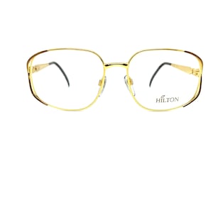 May include: Gold-tone metal eyeglasses with a rectangular frame and clear lenses. The brand name "HILTON" is printed on the right lens.