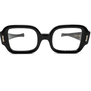 May include: Black square-framed spectacles with a thick, glossy frame. The temples are dark grey with a curved design. Gold-coloured accents are on the outer corners of the frame. The lenses are clear.