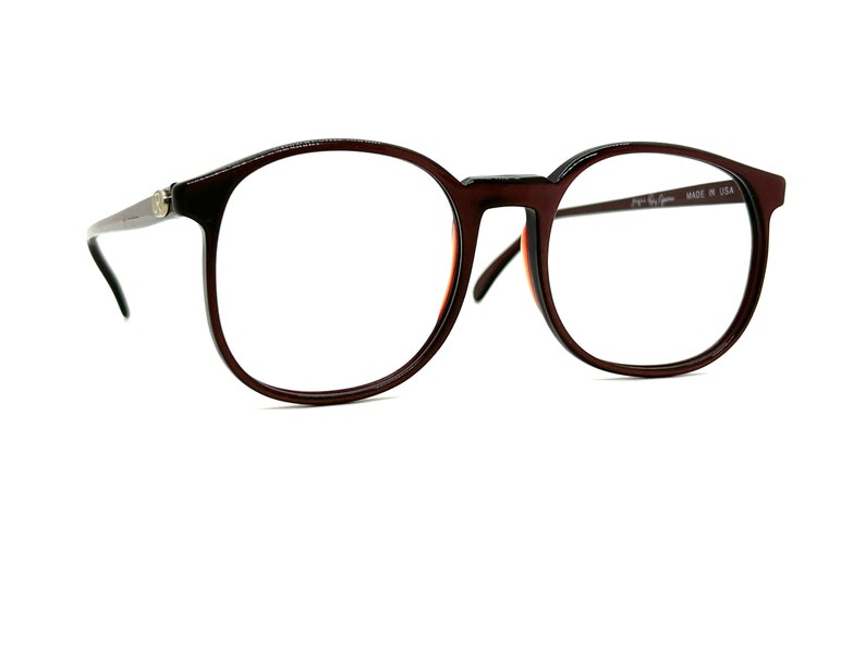 May include: Brown round eyeglasses with a dark brown frame and clear lenses. The glasses have a small gold logo on the left temple and the text "Vogue Eye Wear MADE IN USA" on the right temple.