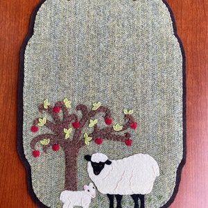 May include: A green felt oval-shaped table runner with a brown felt border. The runner features an embroidered design of a white sheep and a lamb standing under a brown tree with red apples and green leaves.