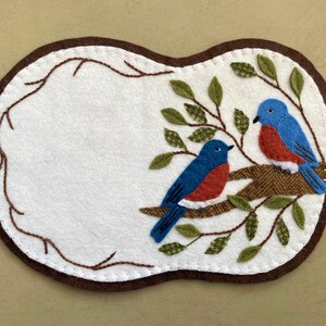 May include: A white felt oval with a brown felt border, featuring two bluebirds with red breasts perched on a brown branch with green leaves. The birds are embroidered with white stitching.