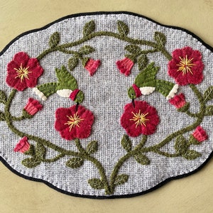 Może przedstawiać: Szary owalny dywan wykonany techniką haftu igłowego z kwiatowym wzorem przedstawiającym dwa zielone kolibry, czerwone kwiaty i zielone liście.
