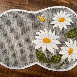 May include: A grey felt coaster with a white border and three white felt daisies with yellow centres. A yellow felt bee is in the top right corner.