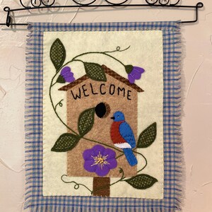 May include: A felt wall hanging with a bluebird perched on a birdhouse. The birdhouse is surrounded by green leaves and purple flowers. The birdhouse has the word "WELCOME" written on it. The hanging is framed with a blue and white plaid border.