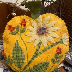 May include: A handcrafted, plush pumpkin decoration in a vibrant yellow hue. The pumpkin is adorned with a large, light yellow sunflower and green leaves, with red berry accents. The stem is green. This decorative item is perfect for fall.