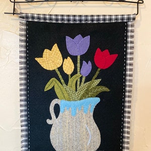 Puede incluir: Una pared de fieltro negro con un borde a cuadros blanco y negro. El centro de la pared presenta un ramo de tulipanes coloridos en una jarra azul y blanca.