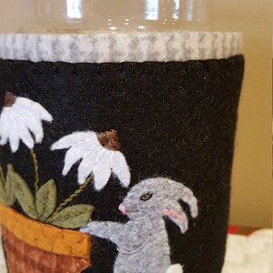 May include: A black felt jar cozy with a white daisy, green leaves, a brown pot, and a gray bunny. The cozy is decorated with a white and gray checkered pattern.