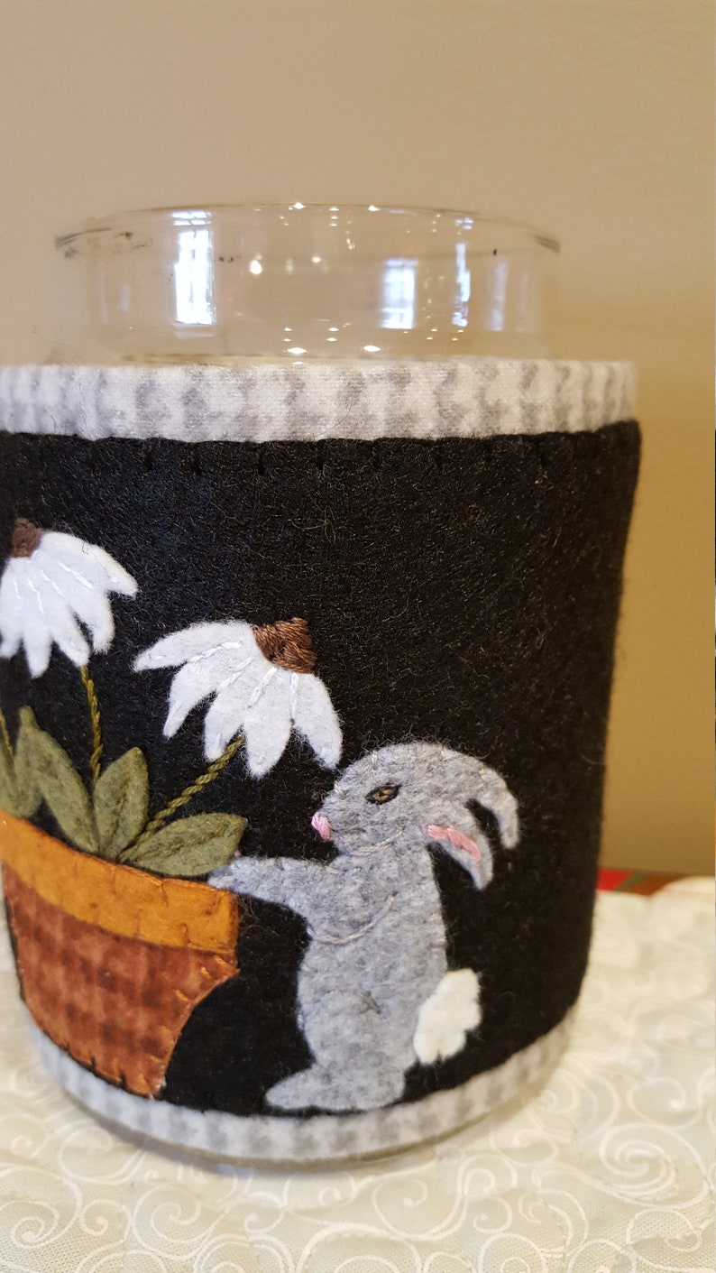 May include: A black felt jar cozy with a white daisy, green leaves, a brown pot, and a gray bunny. The cozy is decorated with a white and gray checkered pattern.