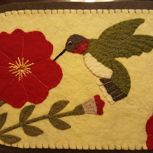 May include: A felt wall hanging with a green and white hummingbird with a red throat, hovering near a red flower with a yellow center. The background is a light yellow.