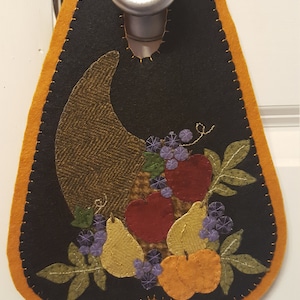 Könnte beinhalten: Ein Filztürhänger in Form eines Füllhorns mit schwarzem Hintergrund und orangefarbenem Rand. Das Füllhorn ist gefüllt mit Filzfrüchten, darunter Äpfel, Birnen und Kürbisse. Es gibt auch Filzblätter und Trauben.