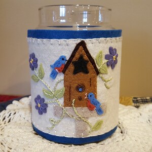 May include: A white felt candle jar cozy with blue trim, featuring an embroidered birdhouse with two bluebirds, green vines, and purple flowers.