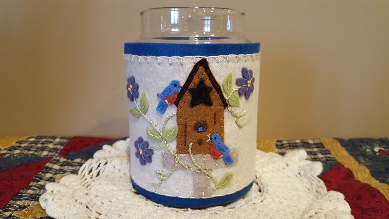 May include: A white felt candle jar cozy with blue trim, featuring an embroidered birdhouse with two bluebirds, green vines, and purple flowers.